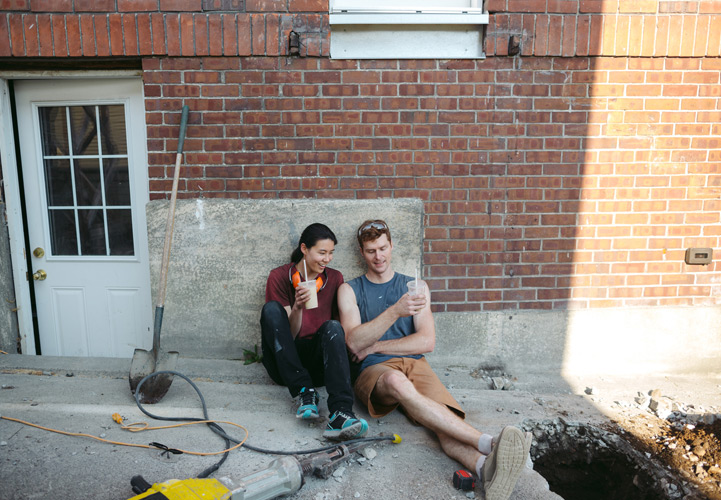 Mann und Frau mit Getränken an Wand sitzend, davor Baugerätschaften