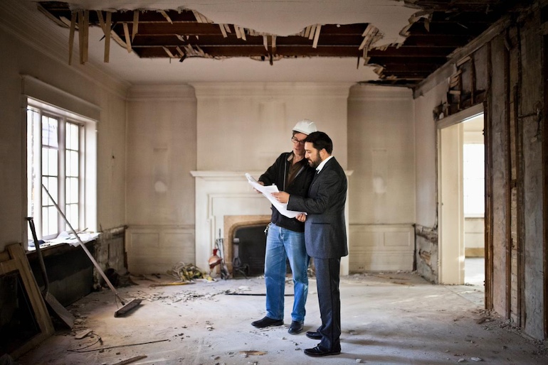 Zwei Männer sehen sich einen Plan an, sie stehen im inneren eines baufälligen Hauses