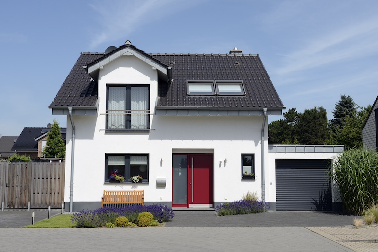 Ein weißes Einfamilienhaus mit roter Tür, Garten und Garage vor blauem Himmel