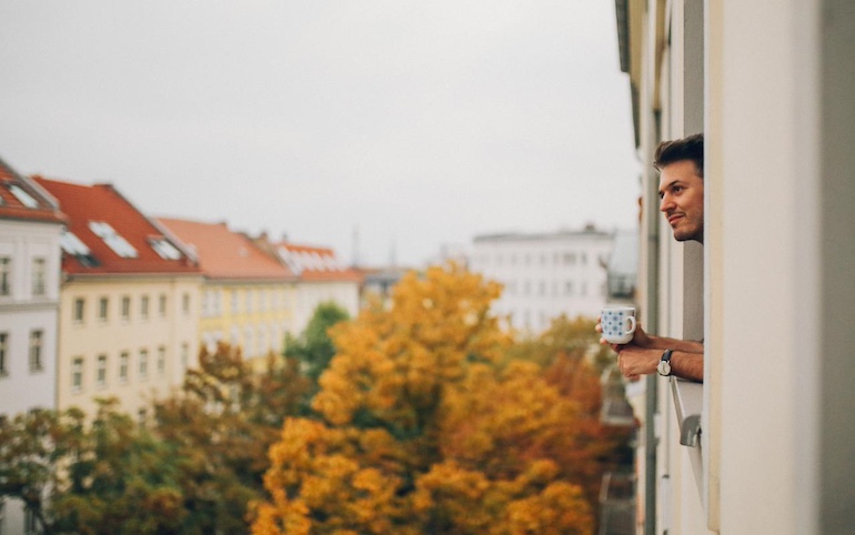 Mann blickt glücklich aus dem Fenster im vierten Stock seiner neuen Wohnung mit Tasse in der Hand