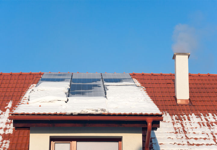 Schneebedeckte Photovoltaikanlage auf einem Dach mit roten Ziegeln, links ein rauchender Schornstein zu sehen