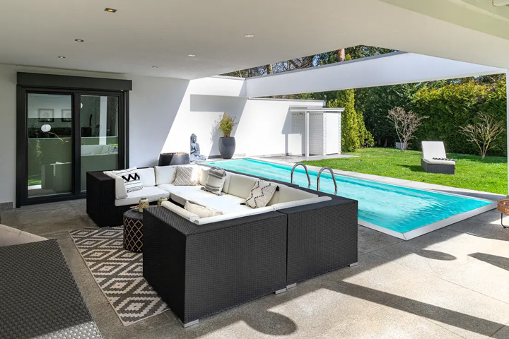 Luxuriöse Poolterrasse einer modernen Villa mit zeitgenössischer Architektur. Die überdachte Terrasse zeigt eine elegante Outdoor-Lounge mit dunklen Rattanmöbeln - eine große, L-förmige Sitzgruppe mit weißen Polstern, ergänzt durch einen niedrigen Couchtisch und einen gemusterten Teppich in geometrischem Design. Der rechteckige Swimmingpool mit kristallklarem, türkisblauem Wasser dominiert den rechten Bildbereich und ist nahtlos in die Terrassenfläche aus hellem Naturstein integriert. Die moderne Architektur des Hauses zeigt große, bodentiefe Glasfronten mit dunklen Rahmen, die eine offene Verbindung zwischen Innen- und Außenbereich schaffen.