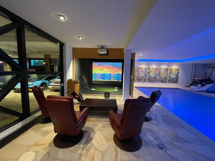 Ein luxuriöser Wellness- und Entertainment-Bereich im Untergeschoss einer modernen Villa mit mehreren Funktionszonen.  Links befindet sich ein Golfsimulator, davor eine hochwertige Sitzgruppe. Rechts im Bild erstreckt sich ein Indoor-Swimmingpool mit türkisblauem Wasser. Rechts ist im Hintergrund durch Glasscheiben eine große Garage mit mehreren Luxusautos zu sehen.