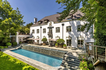 Eine luxuriöse, moderne Villa in zeitgenössischem Architekturstil mit weißer Putzfassade und großzügigen Glasfronten. Das Highlight der Außenanlage ist ein eleganter Swimmingpool mit türkisblauem Wasser, der von einer gepflegten Terrasse aus Naturstein umgeben ist. Der Pool ist von einer niedrigen Natursteinmauer eingefasst und harmonisch in die Gartenlandschaft integriert. Das gesamte Anwesen ist von üppigem Grün umgeben - alte Laubbäume spenden natürlichen Schatten, während gepflegte Rasenflächen und Pflanzbeete eine parkähnliche Atmosphäre schaffen.