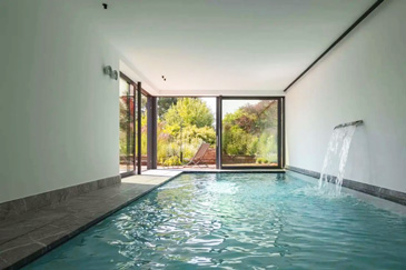 Ein moderner Indoor-Swimmingpool in minimalistischem Design mit direktem Gartenzugang. Der rechteckige Pool mit kristallklarem, türkisblauem Wasser befindet sich in einem hellen, weitläufigen Raum mit weißen Wänden und dunklen Bodenfliesen. Eine elegante Wasserfontäne sprudelt von der rechten Poolseite und schafft eine entspannende Atmosphäre.
