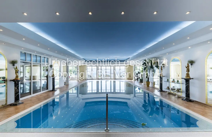 Ein luxuriöser Indoor-Swimmingpool in einem repräsentativen Wellnessbereich mit klassisch-eleganter Architektur. Der rechteckige Pool mit kristallklarem, türkisblauem Wasser dominiert den Raum und ist von einem breiten Rand aus hellem, poliertem Naturstein umgeben. Eine Edelstahl-Poolleiter und ein Handlauf ermöglichen den sicheren Ein- und Ausstieg. Der Poolbereich zeigt aufwendige architektonische Details: hohe Decken mit indirekter LED-Beleuchtung in den Randbereichen, die eine stimmungsvolle Atmosphäre schaffen. Entlang der Wände stehen elegante, dunkle Säulen mit klassischen Kapitellen, die den Raum strukturieren. Die großen, rundbogigen Fenster mit weißen Rahmen lassen natürliches Licht herein und bieten Ausblick ins Grüne.