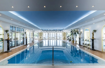 Ein luxuriöser Indoor-Swimmingpool in einem repräsentativen Wellnessbereich mit klassisch-eleganter Architektur. Der rechteckige Pool mit kristallklarem, türkisblauem Wasser dominiert den Raum und ist von einem breiten Rand aus hellem, poliertem Naturstein umgeben. Eine Edelstahl-Poolleiter und ein Handlauf ermöglichen den sicheren Ein- und Ausstieg. Der Poolbereich zeigt aufwendige architektonische Details: hohe Decken mit indirekter LED-Beleuchtung in den Randbereichen, die eine stimmungsvolle Atmosphäre schaffen. Entlang der Wände stehen elegante, dunkle Säulen mit klassischen Kapitellen, die den Raum strukturieren. Die großen, rundbogigen Fenster mit weißen Rahmen lassen natürliches Licht herein und bieten Ausblick ins Grüne.