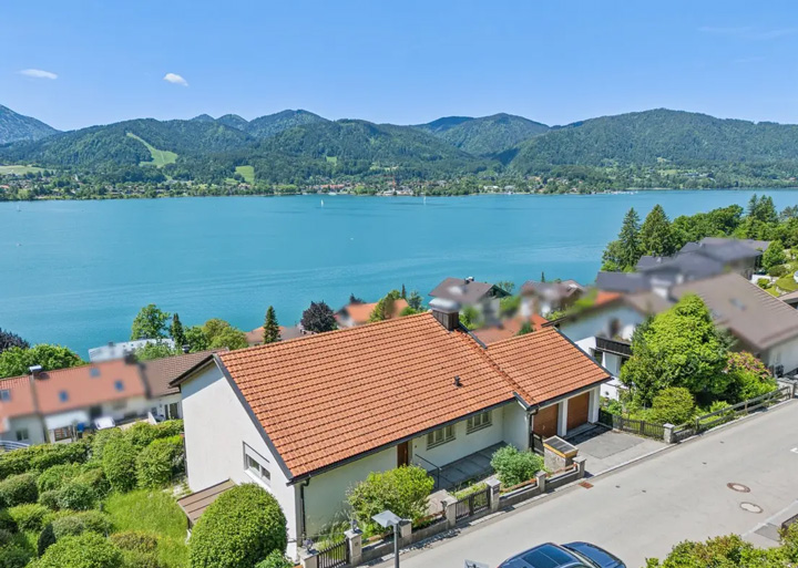 Luftaufnahme einer idyllischen Seelandschaft mit einem türkisblauen See, umgeben von bewaldeten Bergen unter einem klaren blauen Himmel mit wenigen weißen Wolken. Im Vordergrund steht ein Einfamilienhaus mit orangenem Ziegeldach und heller Fassade, das von einem gepflegten Garten umgeben ist. Das Haus liegt in einer Wohnsiedlung am Seeufer, weitere Häuser mit ähnlicher Architektur sind in der Umgebung zu sehen. Eine Straße führt am Haus vorbei, auf der ein blaues Auto geparkt ist. Die grünen Hügel und Berge im Hintergrund sowie die ruhige Wasserfläche des Sees vermitteln eine friedliche, alpine Wohnatmosphäre.