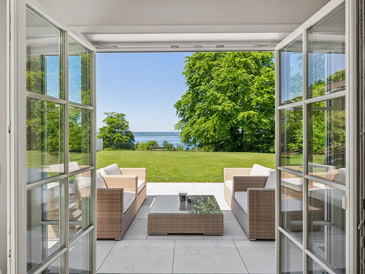 Blick durch eine weit geöffnete, bodentiefe Glasfronttür aus einem Innenraum auf eine moderne Terrasse mit Seeblick. Auf der hellen Steinterrasse steht eine elegante Lounge-Garnitur aus braunem Rattan mit weißen Polstern - bestehend aus zwei Sesseln und einem niedrigen Couchtisch mit Glasplatte. Die Terrasse führt zu einem gepflegten grünen Rasen, der von einem großen, dicht belaubten Baum dominiert wird. Im Hintergrund ist ein blauer See oder Gewässer zu erkennen, umgeben von weiterer grüner Vegetation. Der Himmel ist klar und hellblau.