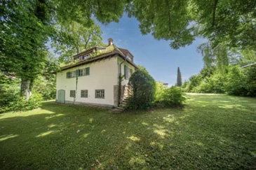 Ein zweistöckiges Landhaus mit heller, cremefarbener Fassade und traditionellem Ziegeldach steht inmitten eines weitläufigen, grünen Gartens. Das Haus verfügt über mehrere Fenster mit grünen Fensterläden und ist teilweise von Efeu bewachsen. Es ist von alten, großen Laubbäumen umgeben, deren Kronen einen natürlichen Rahmen um das Gebäude bilden und für Schatten sorgen. Der gepflegte Rasen erstreckt sich großzügig um das Haus herum. Der blaue Himmel mit vereinzelten Wolken und das warme Sonnenlicht, das durch das Blätterdach fällt, schaffen eine idyllische, ländliche Atmosphäre.