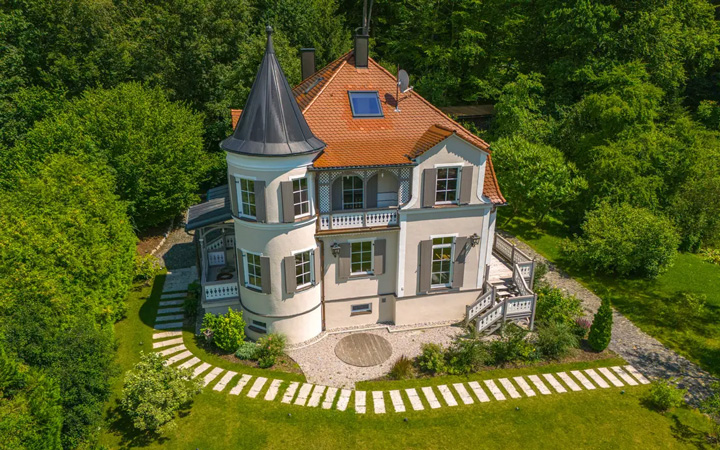 Luftaufnahme einer prächtigen Villa im Jugendstil mit charakteristischem runden Turm und kegelförmigem dunklen Schieferdach. Das zweistöckige Gebäude hat eine hellrosa Fassade mit weißen architektonischen Details und orangeroten Ziegeldächern. Markante Merkmale sind der Rundturm mit Erkerfenstern, mehrere Balkone mit weißen Geländern und eine breite Freitreppe, die zum Haupteingang führt. Das Haus ist vollständig von dichtem, grünen Laubwald umgeben und steht auf einer gepflegten Rasenfläche. Eine geschwungene Zufahrt aus hellen Steinplatten führt elegant zum Gebäude. Die Villa zeigt klassische Merkmale der Gründerzeit-Architektur und liegt in absoluter Alleinlage inmitten der Natur.