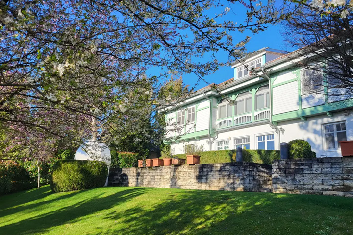 Ein elegantes, mehrstöckiges Wohnhaus mit hellgrüner und weißer Fassade steht hinter einer gepflegten Gartenanlage. Das Gebäude verfügt über mehrere weiße Fenster, Balkone mit weißen Geländern und rote Dachziegel. Im Vordergrund dominiert ein großer Baum mit blühenden Ästen - vermutlich ein Kirsch- oder Apfelbaum in der Frühlingsblüte mit weißen und rosa Blüten. Der Garten ist von einer niedrigen Natursteinmauer eingefasst, auf der mehrere Terrakotta-Töpfe aufgereiht stehen. Eine gepflegte Rasenfläche erstreckt sich vor dem Haus, ergänzt von formgeschnittenen Büschen und Hecken. Der strahlend blaue Himmel und das warme Sonnenlicht, das durch die Baumkrone fällt und Schatten auf den Rasen wirft, schaffen eine frühlingshafte, mediterrane Atmosphäre.