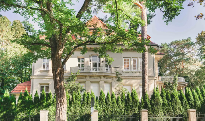Eine elegante Villa im klassizistischen Stil mit weißer Fassade und roten Ziegeldächern, eingebettet in eine parkähnliche Gartenanlage. Das zweistöckige Herrenhaus verfügt über einen repräsentativen Mittelrisalit mit Säulen und einem Balkon im ersten Obergeschoss. Die symmetrische Fassade ist mit weißen Fensterläden und klassischen architektonischen Details geschmückt. Das Gebäude ist von alten, ausladenden Laubbäumen umgeben, die natürlichen Schatten spenden. Im Vordergrund verläuft eine niedrige Hecke aus dicht gepflanzten, immergrünen Koniferen, die das Anwesen zur Straße hin abgrenzt. Steinpfeiler markieren vermutlich den Eingang zum Grundstück. Der bewölkte Himmel und das diffuse Licht verleihen der Szene eine ruhige, gediegene Atmosphäre. Die Villa zeigt typische Merkmale der Gründerzeit-Architektur und vermittelt den Eindruck gehobenen, herrschaftlichen Wohnens in grüner Umgebung.