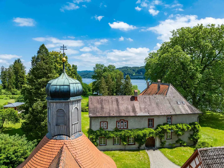 Luftaufnahme eines historischen Herrenhauses. Das stattliche Gebäude verfügt über einen charakteristischen Zwiebelturm mit grüner Patina-Kuppel und goldenem Kreuz, sowie verschiedene Gebäudeteile mit orangeroten Ziegeldächern. Das mehrstöckige Herrenhaus hat eine graue Fassade mit roten Fensterläden und ein großes, rötliches Satteldach. Das herrschaftliche Anwesen ist von saftig grünen Wiesen und alten Laubbäumen umgeben. Im Hintergrund erstreckt sich der blaue Bodensee, umgeben von bewaldeten Hügeln unter einem strahlend blauen Himmel mit weißen Wolken. Die Szene vermittelt eine friedliche, ländliche Atmosphäre und zeigt typische alpine Herrenhaus-Architektur in malerischer Seenlandschaft.