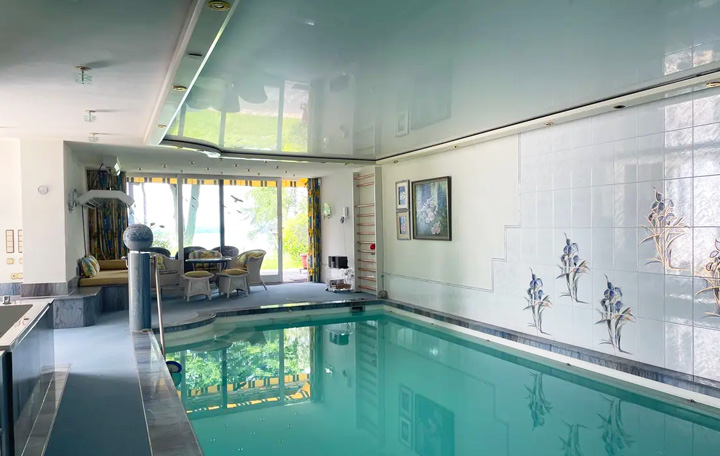 Indoor-Poolbereich mit einem rechteckigen Schwimmbecken mit türkisblauem Wasser. Der Pool ist von hellen Fliesen umgeben und verfügt über eine Edelstahl-Poolleiter. Die Wände sind mit weißen Fliesen verkleidet, die mit floralen Motiven - blauen Iris-Blumen - verziert sind. Die Decke ist in einem glänzenden Grünton gehalten und reflektiert das Licht. Im Hintergrund ist ein gemütlicher Loungebereich mit dunklen Sesseln und einem niedrigen Tisch zu sehen, der durch große Fenster mit Blick ins Grüne natürlich beleuchtet wird. An der Wand hängen gerahmte Bilder, und die Beleuchtung erfolgt durch diskrete Einbaustrahler in der Decke.