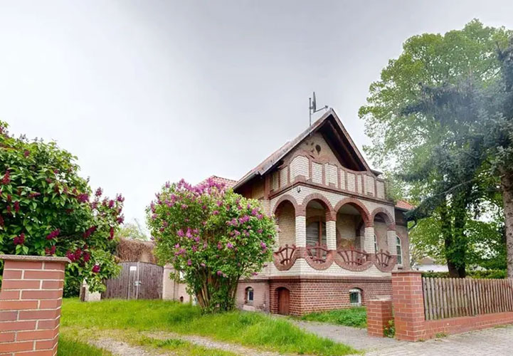 Eine Backsteinvilla im historisierenden Stil mit charakteristischer roter Klinkerfassade und roten Ziegeldächern. Das zweistöckige Gebäude zeigt aufwendige Architekturdetails: Im Obergeschoss befinden sich drei elegante Rundbogenöffnungen mit schmiedeeisernen Balkonen, die von schlanken Säulen getragen werden. Das Erdgeschoss hat ebenfalls Rundbogenfenster und eine zentrale Eingangstür. Die asymmetrische Dachlandschaft mit verschiedenen Giebeln und einem steilen Hauptdach verleiht der Villa einen märchenhaften Charakter. Das Anwesen ist von einer gepflegten Gartenanlage umgeben: Links blüht ein großer Strauch mit violetten Blüten (möglicherweise Flieder), rechts stehen große, grüne Laubbäume. Eine niedrige Backsteinmauer mit Holzzaun grenzt das Grundstück.