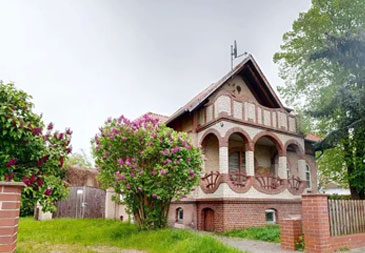 Eine Backsteinvilla im historisierenden Stil mit charakteristischer roter Klinkerfassade und roten Ziegeldächern. Das zweistöckige Gebäude zeigt aufwendige Architekturdetails: Im Obergeschoss befinden sich drei elegante Rundbogenöffnungen mit schmiedeeisernen Balkonen, die von schlanken Säulen getragen werden. Das Erdgeschoss hat ebenfalls Rundbogenfenster und eine zentrale Eingangstür. Die asymmetrische Dachlandschaft mit verschiedenen Giebeln und einem steilen Hauptdach verleiht der Villa einen märchenhaften Charakter. Das Anwesen ist von einer gepflegten Gartenanlage umgeben: Links blüht ein großer Strauch mit violetten Blüten (möglicherweise Flieder), rechts stehen große, grüne Laubbäume. Eine niedrige Backsteinmauer mit Holzzaun grenzt das Grundstück.