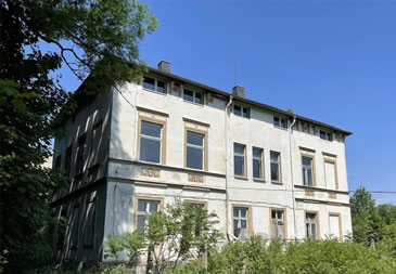 Ein dreistöckiges Gründerzeitgebäude mit klassischer Fassadenarchitektur des späten 19. oder frühen 20. Jahrhunderts. Das Gebäude hat eine verputzte, hellgraue Fassade mit deutlichen Spuren von Witterung und Alter. Die symmetrisch angeordneten Fenster sind mit hellen Sandsteingewänden umrahmt und teilweise mit kleinen Balkonen oder Fensterbänken ausgestattet. Charakteristische architektonische Details sind die horizontalen Gesimsbänder zwischen den Stockwerken und verzierte Fensterbrüstungen.