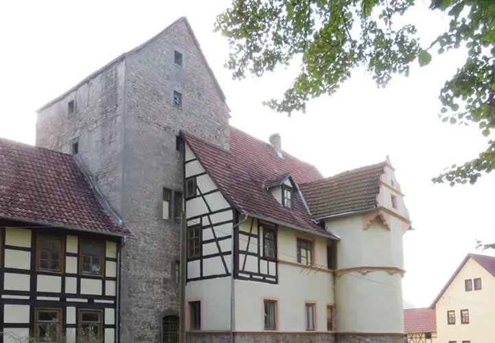 Ein stattliches, weißes Schloss oder Herrenhaus mit einem charakteristischen grünen Kegeldach-Turm. Das mehrstöckige Gebäude hat eine symmetrische, klassizistische Fassade mit regelmäßig angeordneten Fenstern und hellen Fensterläden. Der markante Turm mit seinem grünen Kupferdach ist ein prägendes architektonisches Element. Das Schloss steht in einer parkähnlichen Landschaft mit alten Bäumen unter blauem Himmel.