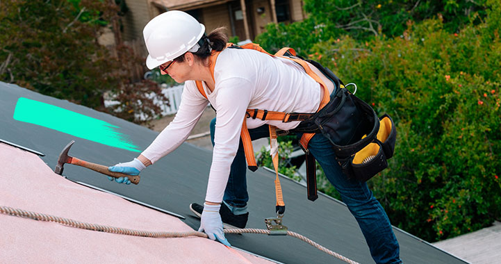 Dachdeckerin mit Werkzeuggürtel und Helm steht auf einem Dach und bearbeitet mit einem Hammer das Dach