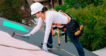 Dachdeckerin mit Werkzeuggürtel und Helm steht auf einem Dach und bearbeitet mit einem Hammer das Dach
