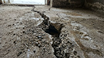 Aufnahme eines massiven strukturellen Schadens in einem Gebäude - ein tiefer, breiter Riss durchzieht den Betonboden eines Raums. Der Spalt ist mehrere Zentimeter breit und offenbar sehr tief, mit zerbröckelten, scharfkantigen Rändern.