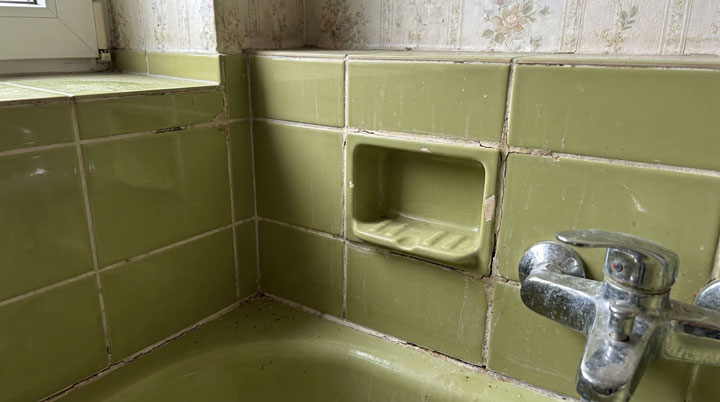 Nahaufnahme einer veralteten Badezimmerausstattung mit deutlichen Anzeichen von Verschleiß und mangelnder Wartung. Das Bild zeigt einen Bereich um ein Waschbecken mit olivgrünen Keramikfliesen aus den 1960er oder 1970er Jahren, die typisch für diese Bauepoche sind.