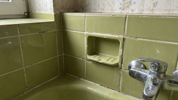 Nahaufnahme einer veralteten Badezimmerausstattung mit deutlichen Anzeichen von Verschleiß und mangelnder Wartung. Das Bild zeigt einen Bereich um ein Waschbecken mit olivgrünen Keramikfliesen aus den 1960er oder 1970er Jahren, die typisch für diese Bauepoche sind.