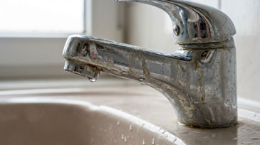 Nahaufnahme eines stark verkalkten und korrodierten Wasserhahns in einem Badezimmer oder einer Küche. Die verchromte Einhebel-Mischbatterie zeigt deutliche Spuren jahrelanger Nutzung und mangelnder Wartung: Die ursprünglich glänzende Chromoberfläche ist großflächig mit weißlichen Kalkablagerungen überzogen, die sich besonders um den Auslauf und an den Verbindungsstellen angesammelt haben.