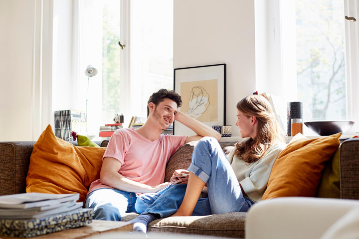Ein junges Paar entspannt gemeinsam auf einem großen, beigen Sofa in einem hellen, modernen Wohnzimmer. Der Mann mit dunklen, welligen Haaren trägt ein rosa T-Shirt und Jeans, während die Frau mit blonden, schulterlangen Haaren einen hellen Pullover und Jeans trägt. Beide sitzen einander zugewandt in entspannter, vertrauter Haltung - er stützt den Kopf auf die Hand, sie hat die Beine angezogen. Ihre Körpersprache und ihr Lächeln vermitteln Intimität und Wohlbefinden. Das Wohnzimmer ist hell und einladend gestaltet: große Fenster mit weißen Rahmen lassen viel natürliches Licht herein und bieten Blick ins Grüne. Das Sofa ist mit warmen, senfgelben Kissen dekoriert. Im Hintergrund sind ein Bücherregal mit Büchern, ein gerahmtes Kunstwerk an der weißen Wand und weitere Wohnaccessoires zu sehen, die eine gemütliche, persönliche Atmosphäre schaffen.