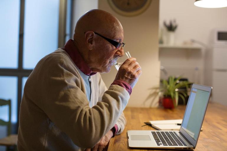 Alter Mann informiert sich über Schonvermögen am Laptop und trinkt ein Glas Wasser