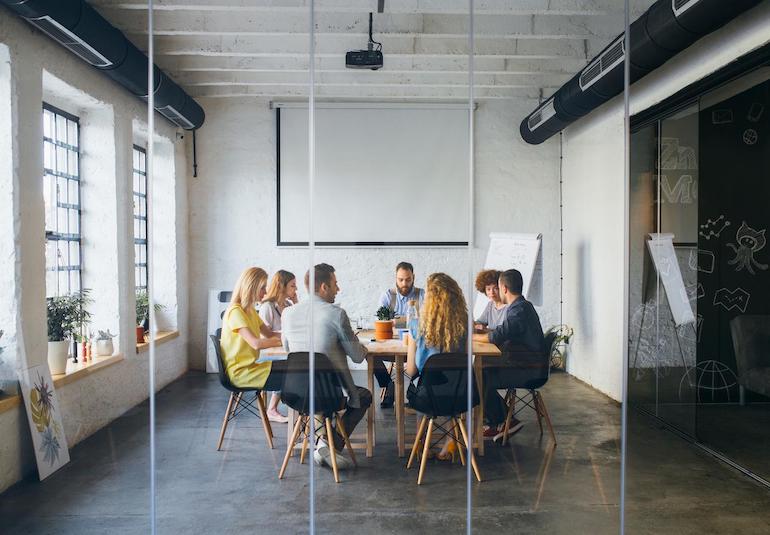 Mehrere Personen sitzen an einem runden Tisch bei einem Meeting, man sieht sie durch eine Glasscheibe
