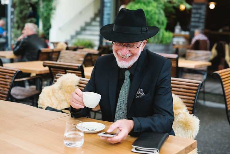 Ein alter Mann mit Hut sitzt draußen in einem gemütlichen Cafè und hält in der einen Hand eine Tasse, während er lächelnd auf sein Smartphone sieht.