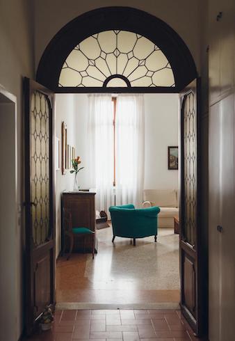 Ein Wohnzimmer mit Holzboden, einem dunkelgrünen Sessel und Möbeln aus dunklem Holz ist durch eine dunkle verzierte Holztür zu sehen, das warme Licht lässt die Szene freundlich wirken