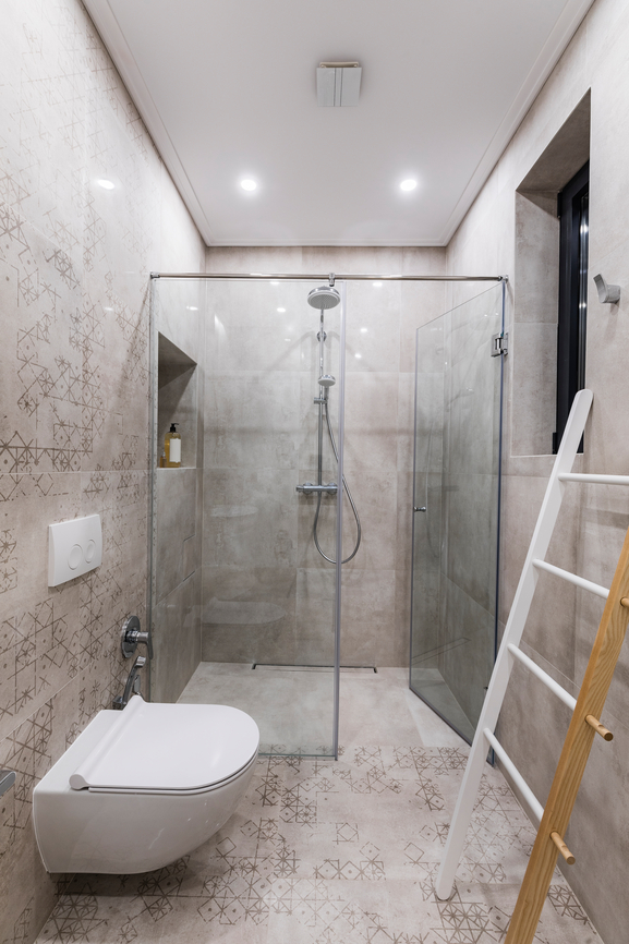 Ein schmales hellgraues Badezimmer mit Toilette links und einer offenen Dusche mit Glastüren am Ende, rechts steht eine Handtuchleiter