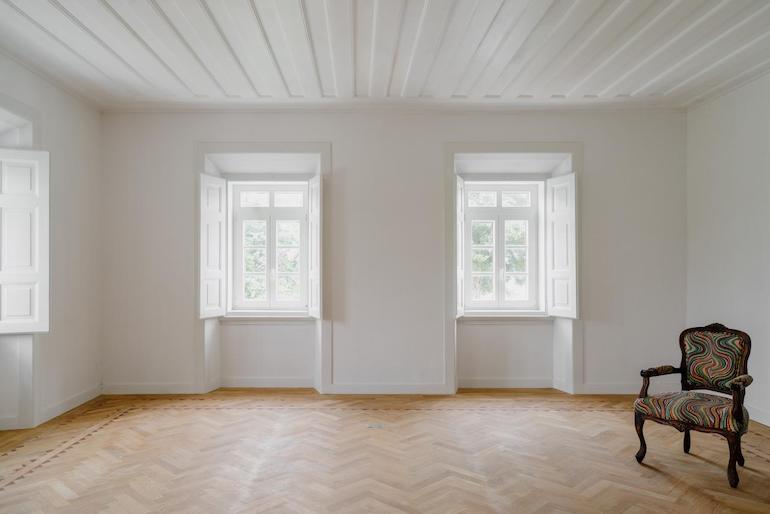 Ein bis auf einen alten gepolsterten Stuhl aus Holz mit Lehnen leerer weißer Raum mit Holzboden, lichtdurchflutet durch zwei Fenster