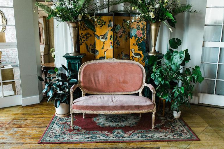 Ein antikes altrosa Sofa steht auf einem alten roten Teppich mit Muster vor Pflanzen und einem alten gelben Paravent mit Vögeln