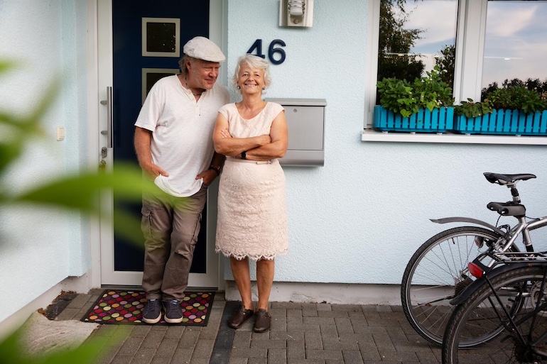 Älteres Paar steht lächelnd vor einem hellblauen Haus mit Pflanzen umrandet.
