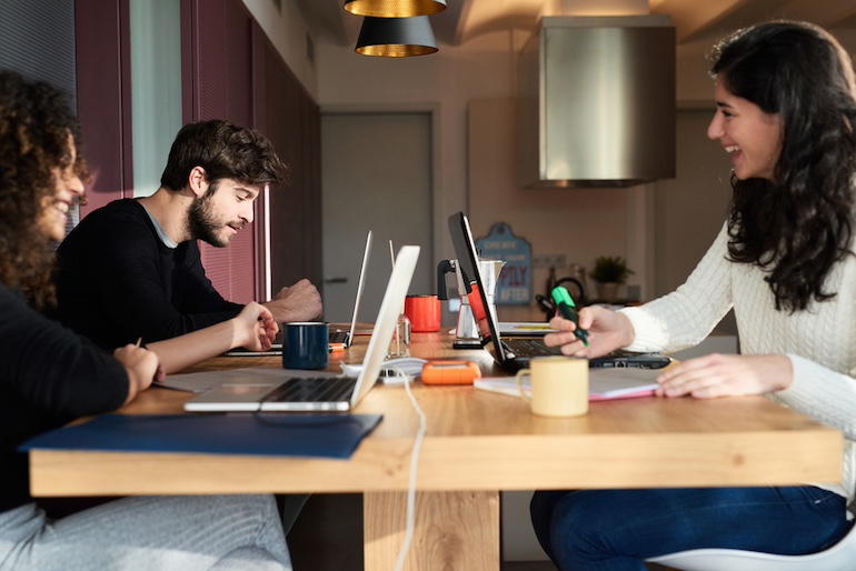 Drei junge Mitglieder einer Wohngemeinschaft lernen an einem Esstisch aus Holz zusammen, mit Textmarkern und Laptops.